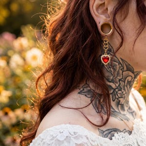 Può includere: Primo piano di un orecchio con un dilatatore dorato e un orecchino pendente. L'orecchino presenta un gioiello rosso a forma di cuore circondato da gemme chiare più piccole. La persona ha lunghi capelli rossi e un tatuaggio floreale.