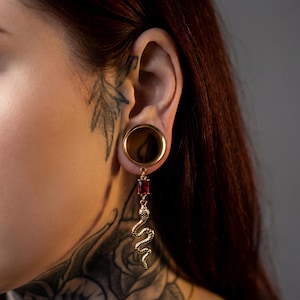 May include: A gold ear gauge with a dangling earring. The earring has a rectangular red gem and a detailed gold snake design. The image is a close-up of the ear and earring.