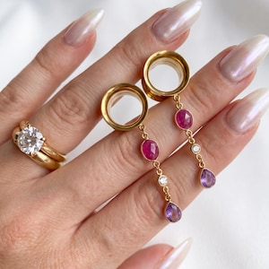 May include: A pair of gold ear tunnels featuring dangling chains with faceted pink oval stones, small clear round stones, and purple pear-shaped stones. A gold ring with a large round clear stone is also visible on the finger.