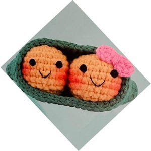 Könnte beinhalten: Gehäkelte Erbsenschote mit zwei lächelnden Erbsen. Die Erbsen sind orange mit rosigen Wangen und schwarzen Augen. Eine Erbse hat eine rosa Schleife. Die Schote ist grün.