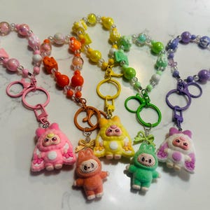 May include: Five keychains with plush figures and beaded wrist straps. The keychains are in pink, orange, yellow, green, and purple. Each keychain has a different plush figure with a cartoon face. The wrist straps are made of beads in matching colors.