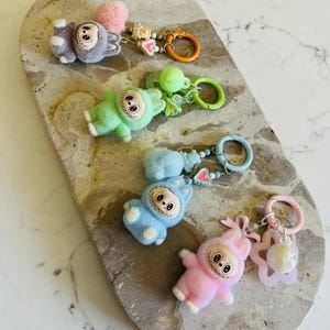 Könnte beinhalten: Eine Sammlung von Plüsch-Schlüsselanhängern in verschiedenen Farben, darunter Grau, Grün, Blau und Rosa. Jeder Schlüsselanhänger zeigt eine Cartoonfigur mit einem lächelnden Gesicht und ist an einem Schlüsselring befestigt. Einige Schlüsselanhänger haben zusätzliche dekorative Elemente wie Pompons und Charms.