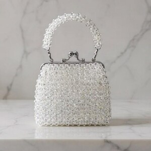 May include: A small, square-shaped handbag covered in clear beads. The bag has a silver-colored metal frame and a beaded handle. The clasp is also silver-colored. The bag is sitting on a white surface.