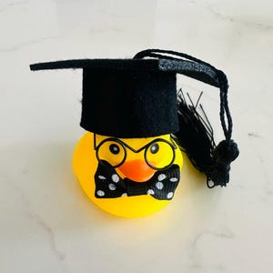 Puede incluir: Un patito de goma amarillo con birrete negro, gafas y pajarita de lunares blancos y negros. El pato tiene pico naranja y está listo para graduarse. Un regalo divertido para un graduado.