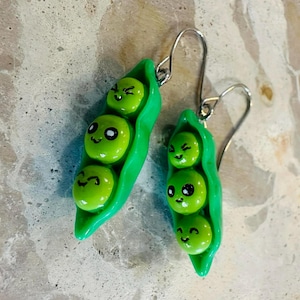 Könnte beinhalten: Ein Paar skurrile Erbsen-Ohrringe. Jeder Ohrring hat drei grüne Erbsen mit lächelnden Gesichtern in einer hellgrünen Schote. Die Ohrringe haben silberfarbene Fischhaken-Ohrdrähte.