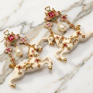 Könnte beinhalten: Ein Paar goldfarbene Ohrhänger mit cremefarbenen Einhornfiguren, die mit kleinen roten und klaren Kristallen verziert sind. Der obere Teil enthält einen quadratischen rosa Kristall, dekorative Metallelemente und eine weiße Perle.
