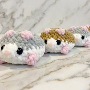 Könnte beinhalten: Eine Reihe kleiner, gehäkelter Plüsch-Hamster aus weichem Garn. Jeder Hamster hat einen weißen Bauch, einen farbigen Rücken in Grau- oder Brauntönen, kleine rosa Pfoten, eine winzige rosa Nase und runde, schwarze Kunststoffaugen als Details.