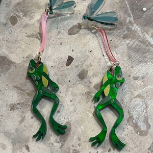 Könnte beinhalten: Ein Paar skurriler Ohrringe mit einem grünen Froschdesign und gelben Akzenten. Die Ohrringe hängen an einem rosa, gebogenen Element, das mit einem blau-weiß gefärbten Libellen-Oberteil verbunden ist. Auf einer strukturierten, grauen Oberfläche.