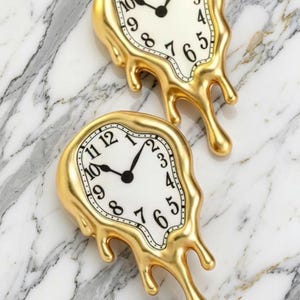 Könnte beinhalten: Zwei Wanduhren mit einer verzerrten, schmelzenden Optik. Jede Uhr verfügt über einen metallisch goldenen Rahmen mit unregelmäßigen, tropfenden Kanten, ein weißes Zifferblatt, schwarze Ziffern und schwarze Zeiger.