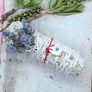 May include: A white sage smudge stick with dried lavender and a small shell with an evil eye design. The sage is wrapped with white string and tied with a red cord. Green sprigs are visible.