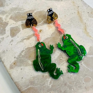 Könnte beinhalten: Ein Paar skurriler Ohrringe mit einem grünen Froschdesign mit roten Augen und einer Fliege oben. Die Frösche sind durch eine rosa, wellenförmige Kette mit der Fliege verbunden. Die Ohrringe sind auf einem neutralen Hintergrund abgebildet.