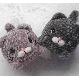 Könnte beinhalten: Zwei handgefertigte Katzenplüschtiere aus Häkelarbeit. Eines ist weichgrau, das andere ist ein gedämpftes Lila. Beide haben schwarze Augen, rosa Nasen und weiße Mäuler. Die Plüschtiere sind aus weichem, strukturiertem Garn gefertigt.