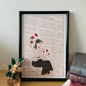 Peut inclure: Illustration encadrée d'une personne lisant dans une bibliothèque, avec le mot « LITERATURE » au-dessus des étagères. L'illustration est en noir et blanc avec des accents floraux rouges. Un petit pot en forme de hérisson avec des fleurs séchées est posé sur une surface en bois.