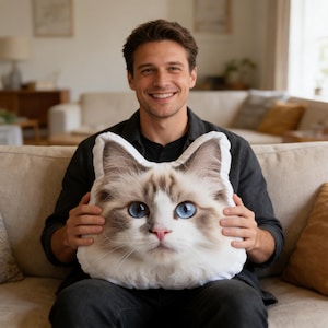 May include: A plush throw pillow shaped like a cat head, featuring a realistic print of a long-haired cat with white and brown fur, bright blue eyes, and a pink nose. The pillow is contoured to the shape of the cat's ears and face.