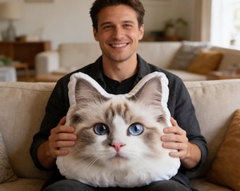 May include: A plush throw pillow shaped like a cat head, featuring a realistic print of a long-haired cat with white and brown fur, bright blue eyes, and a pink nose. The pillow is contoured to the shape of the cat's ears and face.