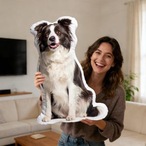 May include: A plush, dog-shaped pillow featuring a Border Collie. The pillow has a white background with a black and white dog design. The dog is sitting with its mouth open. The pillow is being held by a person.