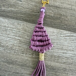 May include: A handmade lavender macrame tassel keyring with a gold star-shaped clasp and a gold-wrapped accent. The tassel has a beaded accent and a fringe of lavender cords.