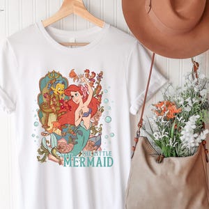 Puede incluir: Camiseta blanca con una ilustraci&oacute;n colorida de Ariel de La Sirenita. El dise&ntilde;o incluye a Ariel, Flounder y elementos florales. El texto "THE LITTLE MERMAID" est&aacute; impreso en turquesa. La camiseta est&aacute; colgada en una percha de madera.