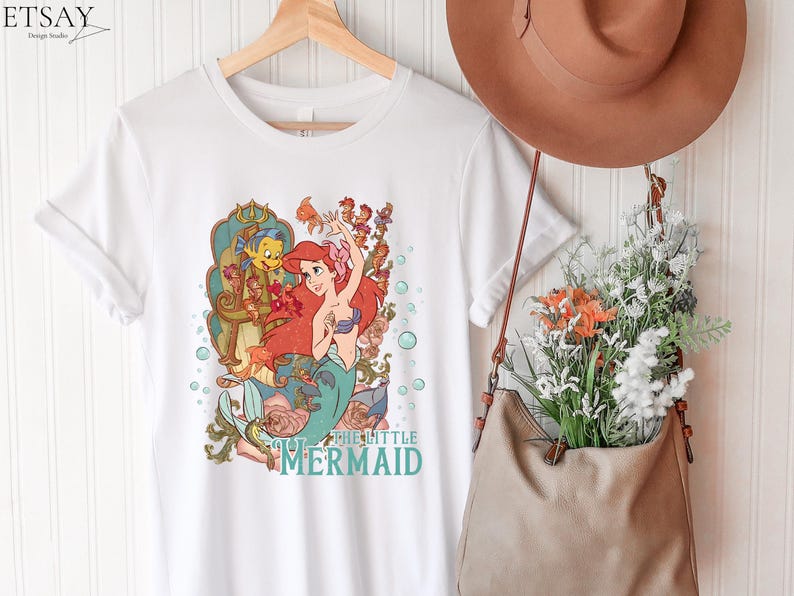 Puede incluir: Camiseta blanca con una ilustraci&oacute;n colorida de Ariel de La Sirenita. El dise&ntilde;o incluye a Ariel, Flounder y elementos florales. El texto "THE LITTLE MERMAID" est&aacute; impreso en turquesa. La camiseta est&aacute; colgada en una percha de madera.