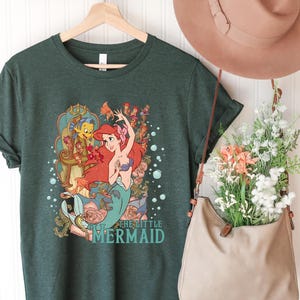 Puede incluir: Una camiseta verde oscuro con un dise&ntilde;o de Ariel de "La Sirenita". El dise&ntilde;o muestra a Ariel con cabello rojo y una cola turquesa, con el texto "The Little Mermaid". La camiseta est&aacute; colgada en una percha de madera.