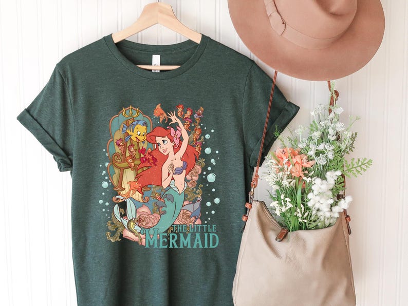 Puede incluir: Una camiseta verde oscuro con un dise&ntilde;o de Ariel de "La Sirenita". El dise&ntilde;o muestra a Ariel con cabello rojo y una cola turquesa, con el texto "The Little Mermaid". La camiseta est&aacute; colgada en una percha de madera.