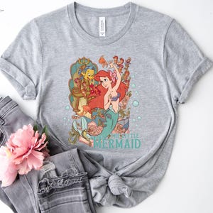 Puede incluir: Una camiseta gris jaspeada con un gr&aacute;fico colorido de Ariel de La Sirenita. El dise&ntilde;o incluye a Ariel, Flounder y Sebasti&aacute;n, con las palabras "THE LITTLE MERMAID" en turquesa. La camiseta se combina con unos vaqueros grises y una flor rosa.