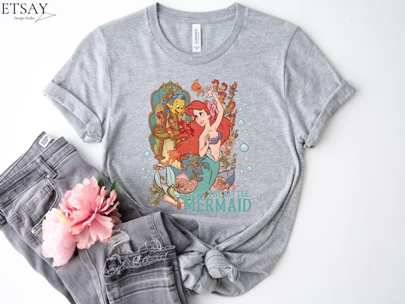 Puede incluir: Una camiseta gris jaspeada con un gr&aacute;fico colorido de Ariel de La Sirenita. El dise&ntilde;o incluye a Ariel, Flounder y Sebasti&aacute;n, con las palabras "THE LITTLE MERMAID" en turquesa. La camiseta se combina con unos vaqueros grises y una flor rosa.