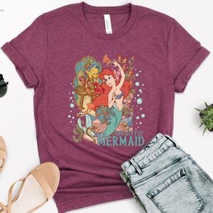 Puede incluir: Camiseta burdeos con un gr&aacute;fico colorido de Ariel de La Sirenita. El dise&ntilde;o incluye a Ariel, Flounder y otras criaturas marinas. El texto "THE LITTLE MERMAID" est&aacute; impreso debajo. La camiseta se combina con vaqueros azules y sandalias.