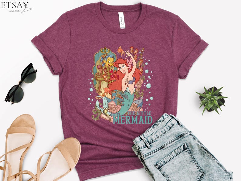 Puede incluir: Camiseta burdeos con un gr&aacute;fico colorido de Ariel de La Sirenita. El dise&ntilde;o incluye a Ariel, Flounder y otras criaturas marinas. El texto "THE LITTLE MERMAID" est&aacute; impreso debajo. La camiseta se combina con vaqueros azules y sandalias.