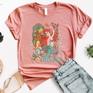 Puede incluir: Una camiseta de color coral con un gr&aacute;fico de Ariel de La Sirenita. El dise&ntilde;o incluye a Ariel con cabello rojo, un sujetador de concha azul y una cola de sirena, junto con otros personajes y el texto "The Little Mermaid".