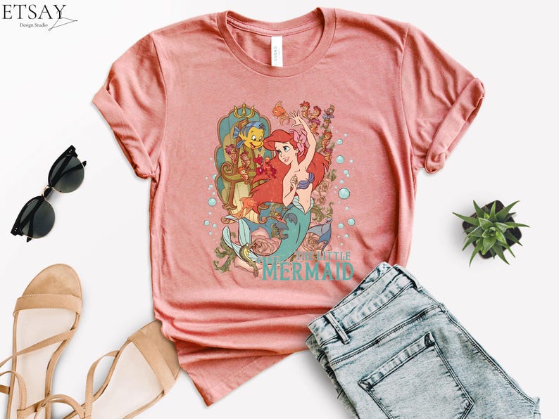 Puede incluir: Una camiseta de color coral con un gr&aacute;fico de Ariel de La Sirenita. El dise&ntilde;o incluye a Ariel con cabello rojo, un sujetador de concha azul y una cola de sirena, junto con otros personajes y el texto "The Little Mermaid".