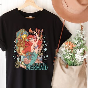 Puede incluir: Camiseta negra con un gr&aacute;fico colorido de Ariel de La Sirenita. El dise&ntilde;o incluye a Ariel, Flounder y otros personajes, con el texto "THE LITTLE MERMAID" en turquesa. La camiseta est&aacute; colgada en una percha de madera.