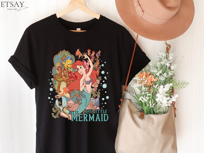 Puede incluir: Camiseta negra con un gr&aacute;fico colorido de Ariel de La Sirenita. El dise&ntilde;o incluye a Ariel, Flounder y otros personajes, con el texto "THE LITTLE MERMAID" en turquesa. La camiseta est&aacute; colgada en una percha de madera.