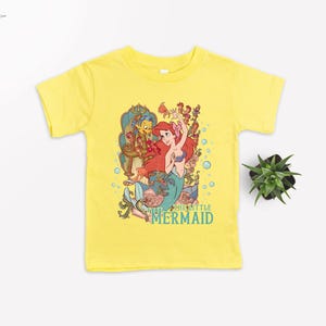 Puede incluir: Camiseta amarilla de manga corta con un gr&aacute;fico colorido de Ariel de La Sirenita. El gr&aacute;fico incluye a Ariel, Flounder, Sebasti&aacute;n y el texto "THE LITTLE MERMAID" en verde azulado. Una peque&ntilde;a planta est&aacute; a la derecha.
