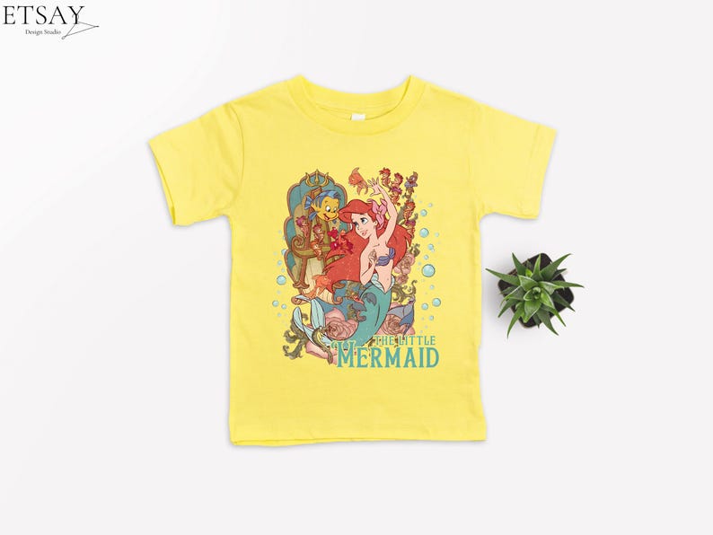 Puede incluir: Camiseta amarilla de manga corta con un gr&aacute;fico colorido de Ariel de La Sirenita. El gr&aacute;fico incluye a Ariel, Flounder, Sebasti&aacute;n y el texto "THE LITTLE MERMAID" en verde azulado. Una peque&ntilde;a planta est&aacute; a la derecha.
