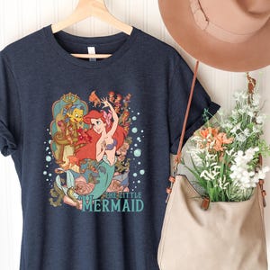 Puede incluir: Camiseta azul marino con un gr&aacute;fico colorido de Ariel de La Sirenita, con el texto "The Little Mermaid". El dise&ntilde;o incluye a Ariel, Flounder y elementos florales. La camiseta est&aacute; colgada en una percha de madera.