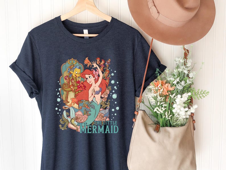 Puede incluir: Camiseta azul marino con un gr&aacute;fico colorido de Ariel de La Sirenita, con el texto "The Little Mermaid". El dise&ntilde;o incluye a Ariel, Flounder y elementos florales. La camiseta est&aacute; colgada en una percha de madera.
