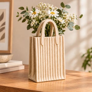 Könnte beinhalten: Eine beige Keramikvase in Form einer Einkaufstasche mit vertikal gerippten Texturen und zwei Henkeln. Die Vase ist mit einem Strauß aus weißen Gänseblümchen und grünen Eukalyptusblättern gefüllt, was ein strukturiertes Blumenarrangement ergibt.