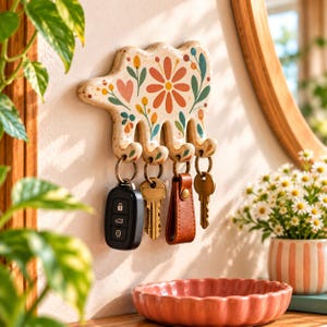 Peut inclure: Un porte-clés mural en céramique couleur crème en forme d'ours avec une finition mouchetée. La surface présente un motif floral peint en orange, jaune et vert. Il possède quatre crochets, chacun portant un anneau métallique avec des clés.