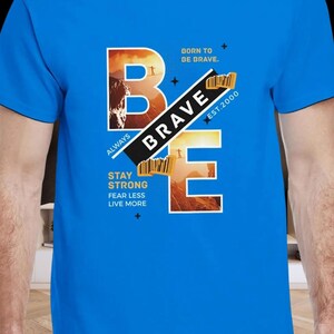 Puede incluir: Una camiseta azul con la palabra "BRAVE" en un diseño audaz. El diseño incluye las letras B y E, con la palabra "BRAVE" en diagonal. El texto adicional incluye "BORN TO BE BRAVE," "ALWAYS," y "STAY STRONG."