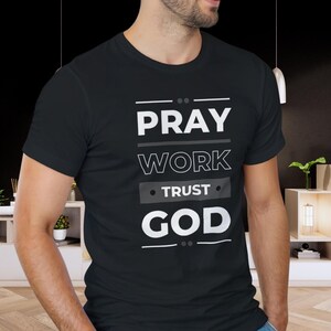 Puede incluir: Camiseta negra con las palabras "PRAY WORK TRUST GOD" en blanco, apiladas verticalmente. El texto está en una fuente limpia, sans-serif, con líneas horizontales encima y debajo de las palabras. La camiseta es de cuello redondo.