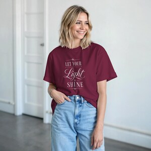 Puede incluir: Camiseta burdeos con la frase "LET YOUR Light SHINE" en blanco. La camiseta se combina con vaqueros azul claro. La persona sonríe. El fondo incluye una puerta blanca.