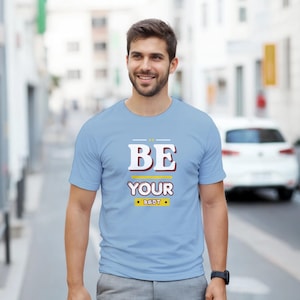 Puede incluir: Una camiseta azul claro con el texto "BE YOUR BEST" en blanco y amarillo. La camiseta es de manga corta y la lleva una persona. El fondo es una escena callejera borrosa con un coche blanco.