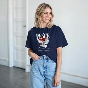 Puede incluir: Camiseta azul marino con la palabra "PEACE" en blanco sobre un gráfico de paloma blanca con una rosa roja y texto debajo. La camiseta se combina con vaqueros azul claro.