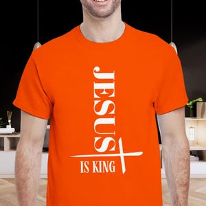 Puede incluir: Camiseta naranja brillante con el texto blanco "JESUS IS KING". La palabra "JESUS" está alineada verticalmente, con una cruz debajo. La camiseta es de cuello redondo.