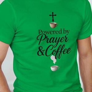 Puede incluir: Camiseta verde con el texto "Powered by Prayer & Coffee" en escritura negra. El diseño incluye una cruz, tazas de café y gráficos de vapor. La camiseta está hecha de un material suave.