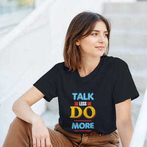 Puede incluir: Camiseta negra de cuello redondo con el texto "TALK LESS DO MORE" en un diseño colorido. El texto es una mezcla de azul, rojo y amarillo. La camiseta la lleva una persona sentada en unos escalones.