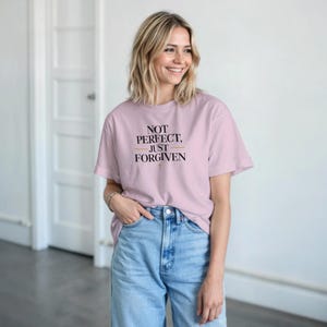 Puede incluir: Una camiseta rosa claro con el texto "NOT PERFECT, JUST FORGIVEN" en letras negras y doradas. La camiseta se combina con unos vaqueros azules. El estilo es informal y cómodo.