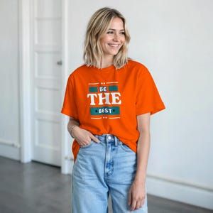 Puede incluir: Camiseta naranja con el texto "BE THE BEST" en blanco, turquesa y naranja. La camiseta es de cuello redondo y se combina con vaqueros azul claro. El look general es informal y cómodo.