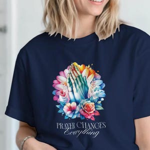 Puede incluir: Camiseta azul marino con un diseño floral colorido y manos rezando. El diseño incluye flores en tonos rosa, azul, amarillo y naranja. El texto "PRAYER CHANGES Everything" está impreso debajo.